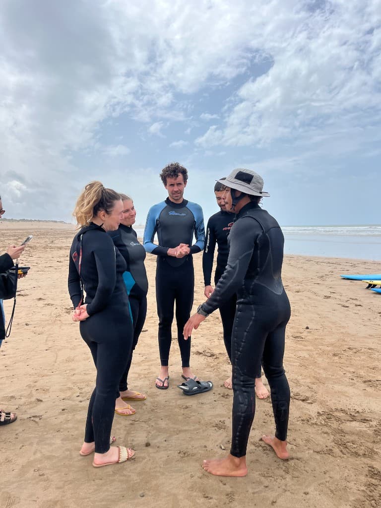 Surf coach briefing a small group in wetsuits in a circle on Essaouira beach — ocean and boards in the background