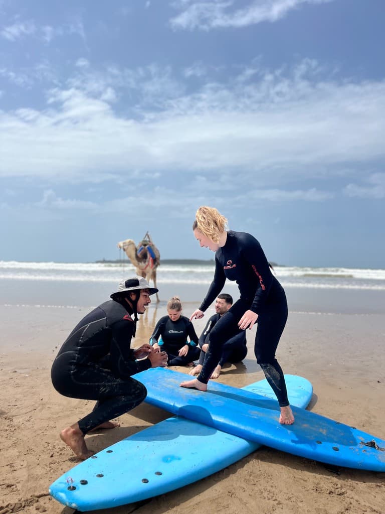 Surf coach correcting stance on stacked boards in Essaouira — students watching on the sand, camel on the shoreline