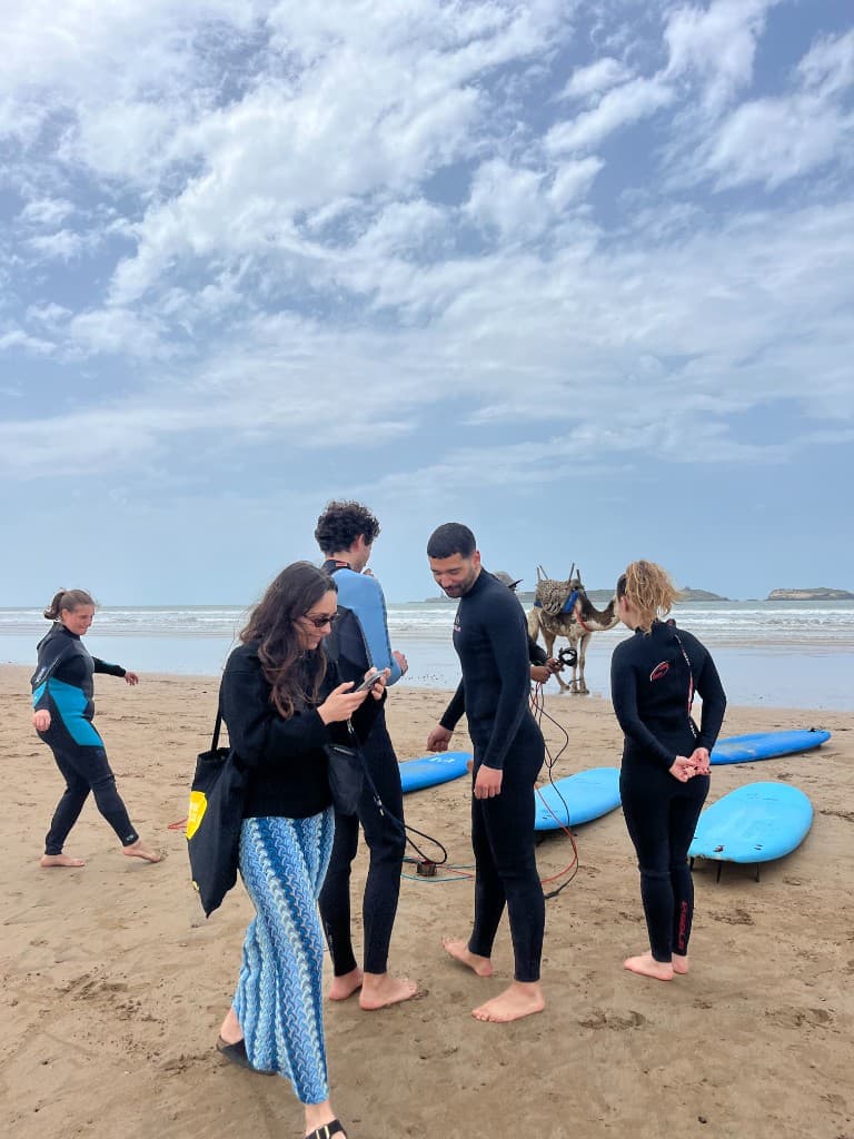 Surf school group on Essaouira beach with blue boards — wetsuits, camel near the shore, and calm Atlantic surf